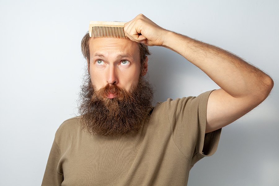 Portrait of adult bald male holding hairbrush, suffering alopecia, looking up with confused facial expression, bearded man wearing T-shirt